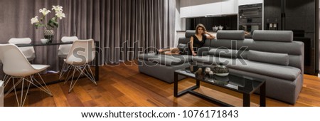 Woman lying on gray sofa in modern apartment with dining table, chairs and kitchenette, panorama