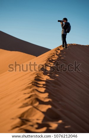 Young male traveler and photographer standing on the top of sand dune photographing sunrise or sunset in desert of Namibia, Africa. Travel photography concept