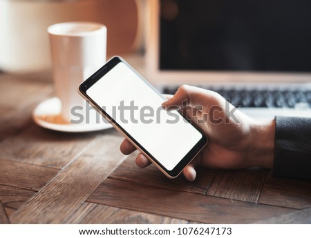 Young man with tattoo holding mobile phone and working on laptop in co-working. Mans hand and phone are on foreground and on focus. Coffee cup and laptop are on background and blurred.