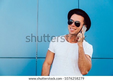 Summer photo of glad young man calling with mobile phone, wearing black hat and sunglasses. Dressed in t-shirt. Outdoors.