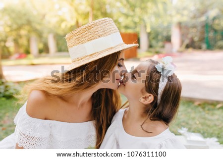 Long-haired young woman wears vintage hat with white ribbon cute kissing daughter posing on nature background. Outdoor portrait of lovely lady and little girl in similar dresses enjoying vacation.