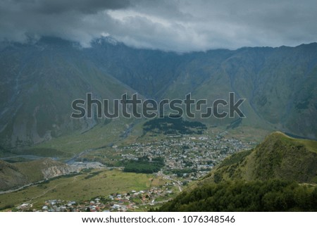 Beautiful landscape with view from the hill on small village in valley and majestic mountains in front under the dramatic clouds and sky.