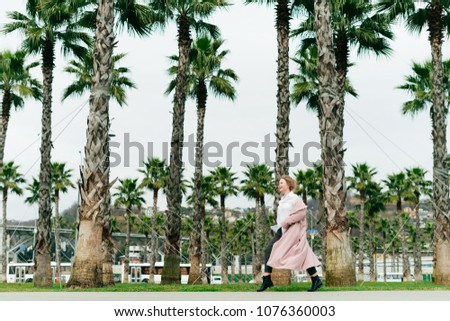 stylish girl in a pink coat walks in the park on a background of tall green palms, enjoying the long-awaited spring