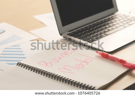 Opened notebook page with word business model with blank screen laptop, pink marker pen and business documents on wooden table in office. Business plan, idea and project model concept.