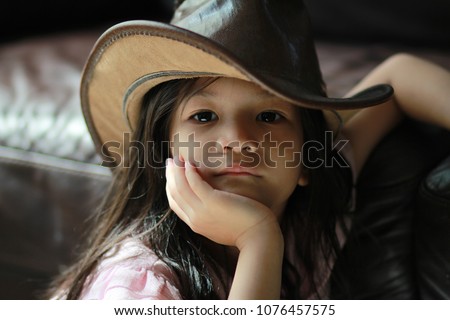 5 year old Asian girl wearing cowboy hat and looking to the camera.Beautiful young lady raise her face on hand while lean to leather sofa.Light touched her cheek.brown color tone.