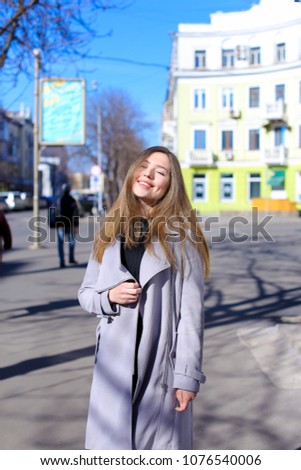 Young lady strolling outdoors near buildings and wearing grey coat with black dress. Concept of walking in spring city.