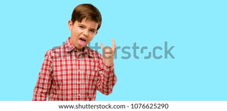Handsome toddler child with green eyes making rock symbol with hands, shouting and celebrating with tongue out over blue background