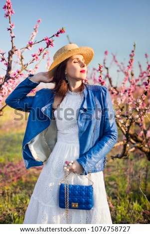 beautiful caucasian girl in white dress and in blue jacket and hat closing eyes under sun lights in flowering garden