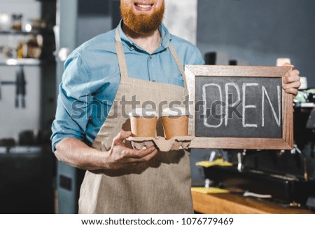cropped shot of young male barista holding chalkboard with lettering open and two papers cups of coffee 