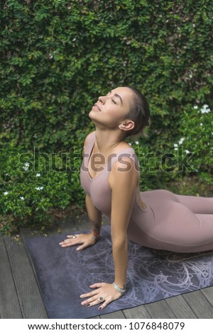 joyful young woman practicing yoga in Upward-Facing Dog (Urdhva Mukha Svanasana) pose in front of wall covered with green leaves