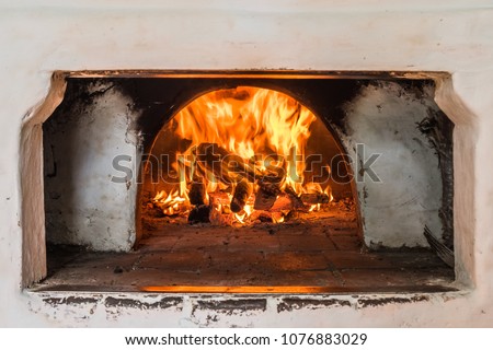 Russian oven painted white with dirty smoked spots. Burning wood on fireplace. Masonry stove used for cooking and domestic heating in traditional Russian, Ukrainian and Belarusian country houses. Royalty-Free Stock Photo #1076883029