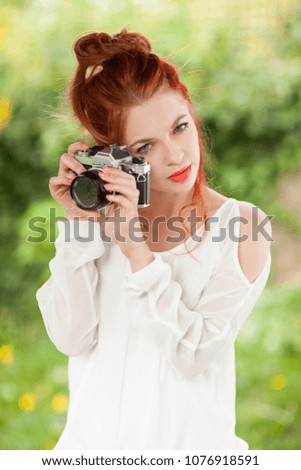 Beautiful young woman with red hair sitting in the garden taking pictures with old, analog camera