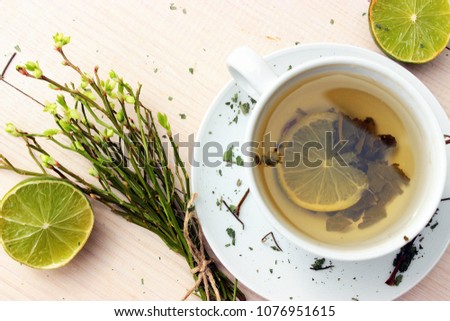 Cup of fresh natural aromatic green tea decorated with dry leafs and small bud of rose, lime and cute bunch of wild flower on white wooden background. Concept for healthy food. Bright green color