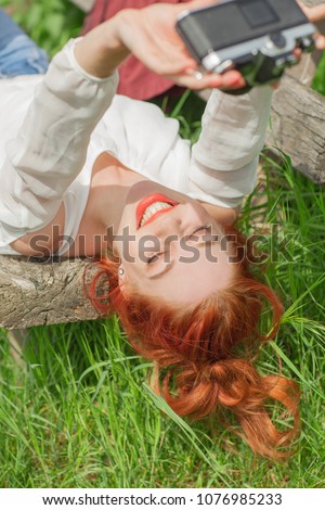 Beautiful young woman relaxing with camera in her hand on garden bench in green grass doing some selfies