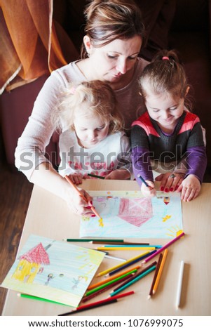 Mom with little girls drawing a colorful pictures of house and playing children using pencil crayons sitting at table indoors. Shot from above