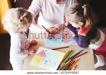 Mom with little girls drawing a colorful pictures of house and playing children using pencil crayons standing at table indoors. Shot from above