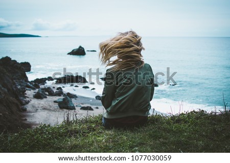 girl sitting on a cliff overlooking the ocean with her hair blowing Royalty-Free Stock Photo #1077030059