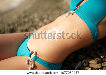 Sunscreen lotion drawn on tanned woman belly skin as sun shape. Suntan cream applied on body. Suncream on stomach. Woman wearing bikini swimsuit sunbathing on the beach. Skin care health concept.