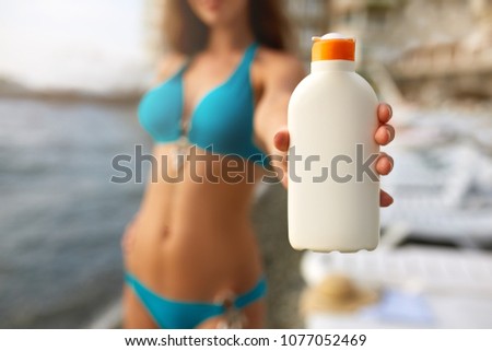 Happy woman shows the suntan or sunscreen cream white bottle over the blurred beach background. Tanned slim girl wearing blue bikini swimsuit and holds sun lotion. White bottle with copy space.