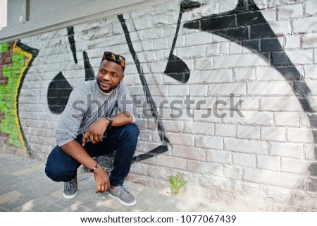 Stylish african american boy on gray sweater and black sunglasses posed at street. Fashionable black guy against graffity wall.