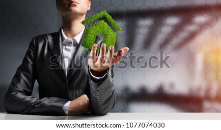 Businessman in suit presenting green plant in form of house symbol with office view and sunlight on background.