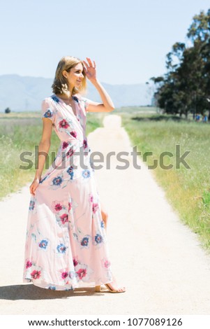 Attractive young blonde Caucasian woman in flowing pink floral print dress posing outside runway style on empty dirt road