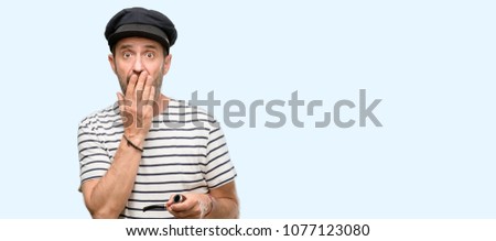 Sailor captain man smoking a tobacco pipe covers mouth in shock, looks shy, expressing silence and mistake concepts, scared isolated over blue background