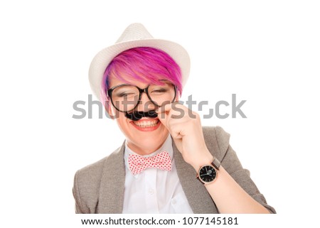 Wonderful millennial model wearing stylish informal outfit, pink hairstyle applying black mustache while laughing happily on white background. Do I look like a gentleman, do mustache fits me concept.
