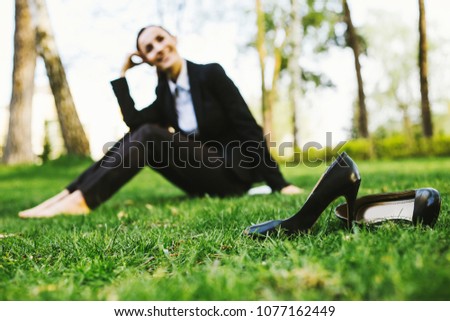 Smiling beautiful young business woman in a black suit sitting on a green lawn in a city park and relax after work
