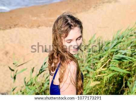 Beautiful blonde in a blue bathing suit on a background of green reeds and the sea. The girl looks down