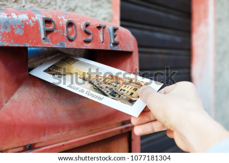 Male hand is drops a postcard in a red postbox in Venice, Italy. The postcard shows a Venice canal.