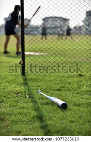 A blue softball bat left out on the out field, waiting for a next batter picking it up, while the batter is ready to hit a pitching ball on the field with a clear sky in summer time.