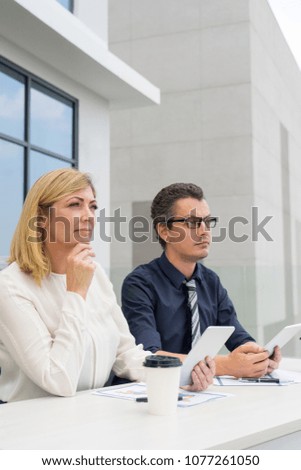 Pensive male and female colleagues working in outdoor cafe. Businesspeople sitting at table with building in background. Outdoor business workplace concept.