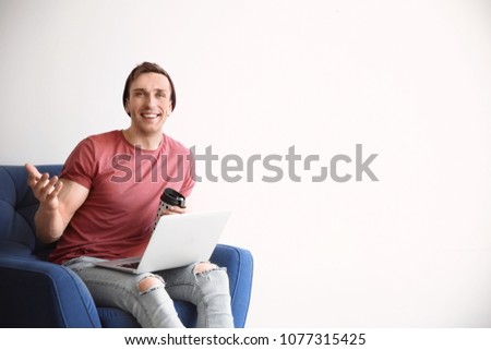 Young blogger with laptop sitting in armchair against light wall