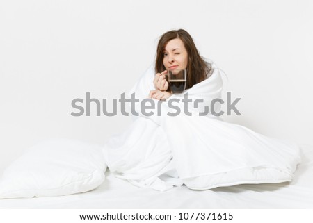 Young tired brunette woman sitting in bed with cup of coffee, white sheet, pillow, wrapping in blanket on white background. Female spending time in room. Rest, relax, good mood concept. Copy space