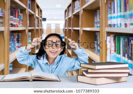 Picture of a happy schoolgirl raising her hands while sitting with books on the table. Shot in the library