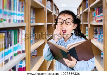 Picture of little girl wearing glasses and holding a textbook while shouting in the library
