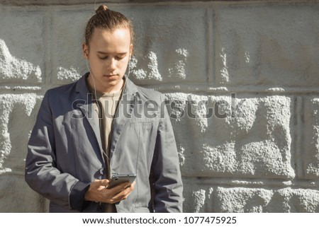 A young man listens to music from a phone on a city street. A pretty boy teenager uses a gadget. A student near the college.
