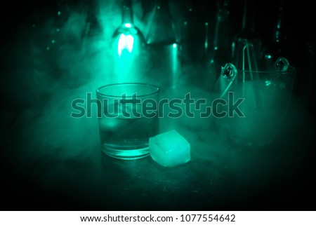 Glass of whisky on wooden bar closeup with bottles blurred view on dark background with light and smoke. Single glass of whisky on ice with a reflective wooden surface. Selective focus. Ready to serve
