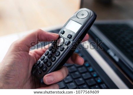 Hand holding a black cordless telephone with a laptop at the background