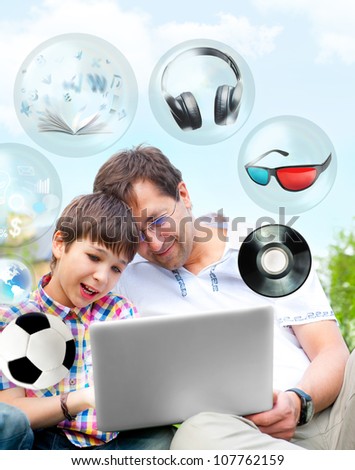 Closeup portrait of happy family: father and his son using laptop outdoor at their backyard sitting on the grass together. Different objects flying from the screen