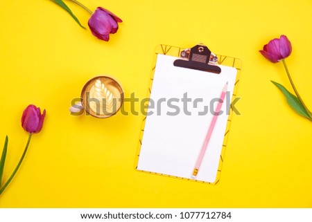Feminine desktop composition with blank sheet clipboard, pencil, coffee cup, tulips bouquet on yellow background. Girlie workplace, flowers for mother's day. Top view, flat lay, close up, copy space.