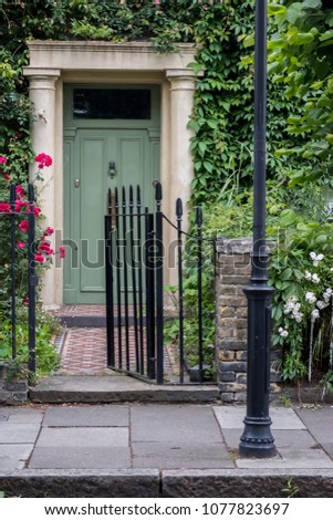 Front door of a British house with white stone frame and surrounded by lush vegetation some red flowers and sidewalk with street lamp in foreground
