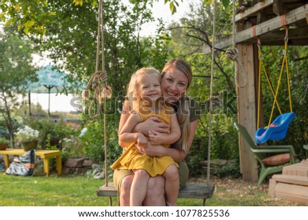 Attractive young mom and daughter on a swing playing in the backyard on a summer day Royalty-Free Stock Photo #1077825536