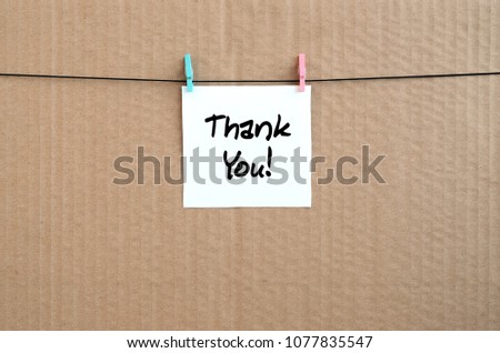 Thank you Note is written on a white sticker that hangs with a clothespin on a rope on a background of brown cardboard