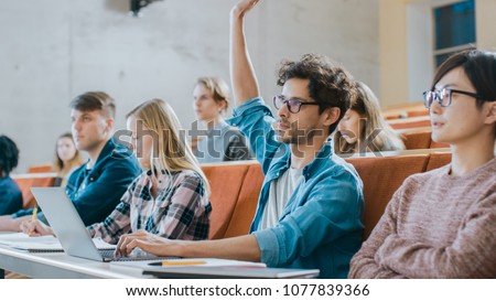 Handsome Hispanic Student Uses Laptop while Listening to a Lecture at the University, He Raises Hand and Asks Lecturer a Question. Multi Ethnic Group of Modern Bright Students. Royalty-Free Stock Photo #1077839366