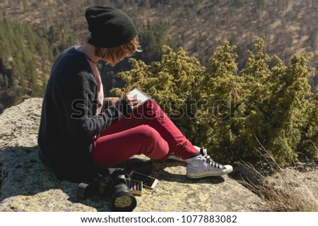 Portrait of a hipster designer photographer girl in hat and sunglasses draws a pastel in her notebook sitting on a rock outdoor in the mountains. The concept of a free freelancer artist