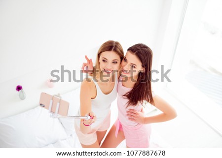 Top view of pretty, charming, cheerful, cute, attractive, joyful girls standing on bed shooting selfie on front camera, using selfish stick and smart phone, hugging, embracing, showing peace symbol
