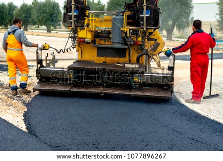 Workers regulate tracked paver laying asphalt heated to temperatures above 160 Â° pavement on a runway Royalty-Free Stock Photo #1077896267