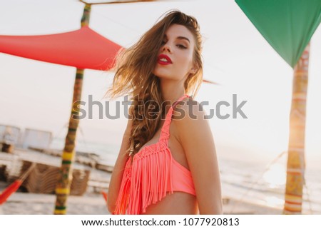 Gorgeous curly girl with red lips looking down while posing at sea beach. Outdoor portrait of spectacular blonde woman wears pink bikini in summer vacation at resort.